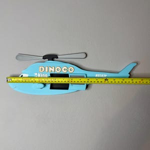 Puede incluir: Una decoraci&oacute;n de pared con forma de helic&oacute;ptero azul claro con el texto DINOCO, The King y N9747P. Una cinta m&eacute;trica amarilla est&aacute; colocada sobre el helic&oacute;ptero, mostrando una longitud de unos 42 cm desde el morro hasta la cola.