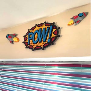 Puede incluir: Decoraci&oacute;n mural estilo c&oacute;mic con una burbuja de di&aacute;logo central "POW!" en azul y amarillo, flanqueada por dos decoraciones de cohetes grises y rojos con llamas amarillas. Los art&iacute;culos est&aacute;n montados en una pared beige.