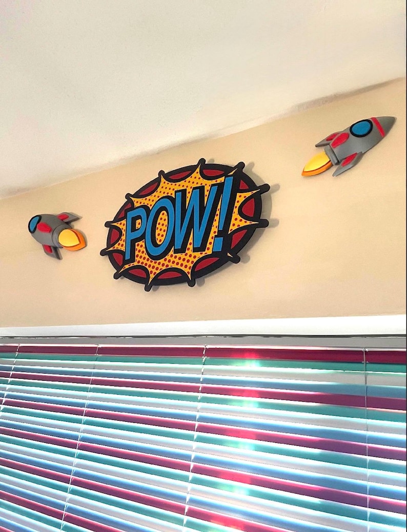 Puede incluir: Decoraci&oacute;n mural estilo c&oacute;mic con una burbuja de di&aacute;logo central "POW!" en azul y amarillo, flanqueada por dos decoraciones de cohetes grises y rojos con llamas amarillas. Los art&iacute;culos est&aacute;n montados en una pared beige.