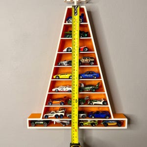 May include: A white and orange triangular shelf unit filled with toy cars. The shelf is approximately 114 cm tall. The unit is mounted on a grey wall and is designed to display a collection of miniature vehicles.