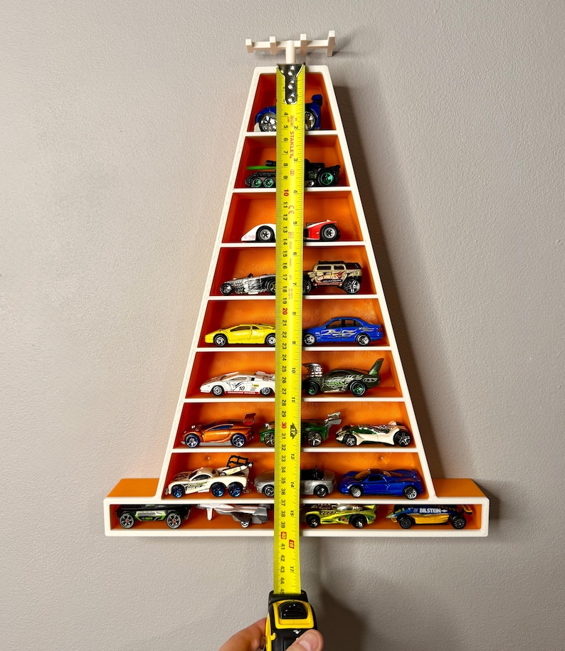 May include: A white and orange triangular shelf unit filled with toy cars. The shelf is approximately 114 cm tall. The unit is mounted on a grey wall and is designed to display a collection of miniature vehicles.