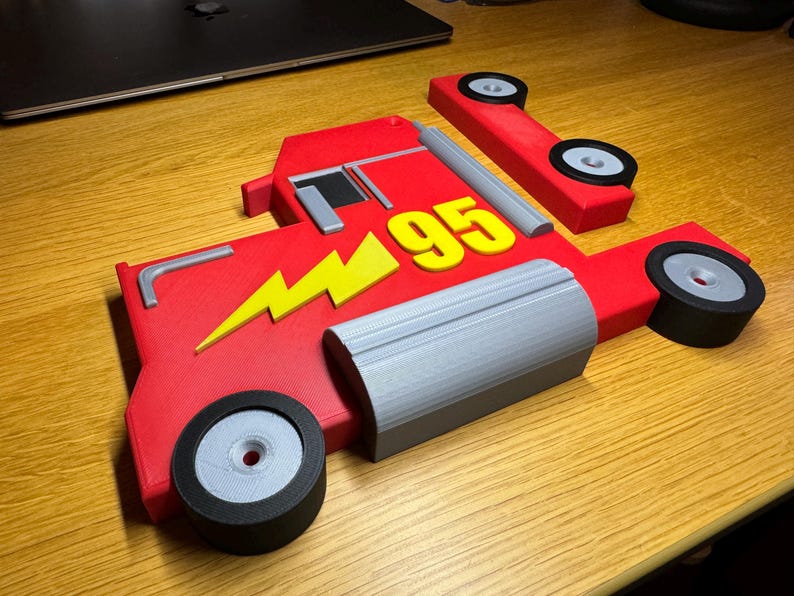 A 3D-printed red truck model inspired by Mack from Cars, featuring a yellow lightning bolt and the number 95. The parts are laid flat, showing two wheels, grey detailing, and black tires, all on a wooden surface.