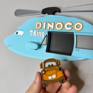 Puede incluir: Un soporte de pared con forma de helic&oacute;ptero azul claro que presenta el texto DINOCO y THE KING en letras blancas y naranjas, con un rotor negro en la parte superior. Debajo hay un peque&ntilde;o personaje de gr&uacute;a marr&oacute;n con ojos verdes y dientes blancos.
