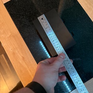 Puede incluir: Un objeto rectangular negro se mide con una regla met&aacute;lica. La regla muestra medidas en cent&iacute;metros, con el objeto de aproximadamente 15 cm de largo. El objeto est&aacute; sobre una superficie negra.