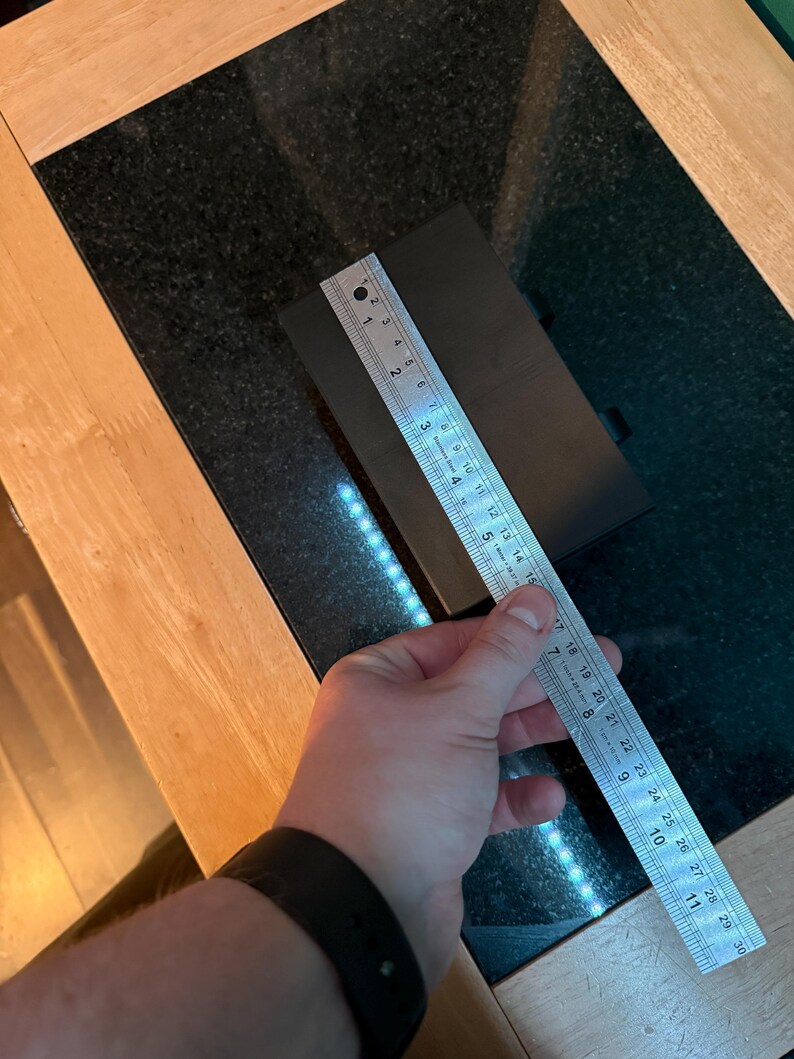 Puede incluir: Un objeto rectangular negro se mide con una regla met&aacute;lica. La regla muestra medidas en cent&iacute;metros, con el objeto de aproximadamente 15 cm de largo. El objeto est&aacute; sobre una superficie negra.