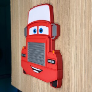 May include: A red cartoon truck wall decor with a white windshield, blue eyes, and a smiling mouth. The truck has a gray grill and gray side mirrors. The decor is mounted on a light brown wooden surface.