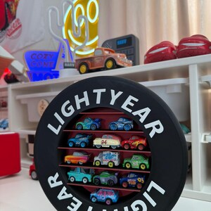 May include: A black, circular display case with the word "LIGHTYEAR" in white lettering, holding a collection of colorful toy cars. The cars are arranged on shelves within the case, showcasing various designs and colors.