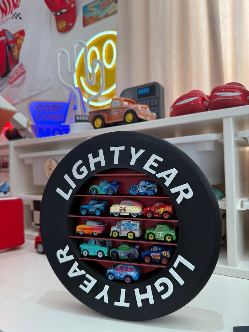 May include: A black, circular display case with the word "LIGHTYEAR" in white lettering, holding a collection of colorful toy cars. The cars are arranged on shelves within the case, showcasing various designs and colors.