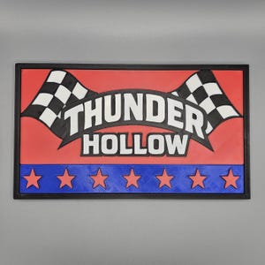 May include: Rectangular sign featuring the text "THUNDER HOLLOW" in white. The sign has a red background with two black and white checkered flags. A blue border with six red stars is at the bottom, all within a black frame.