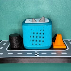 May include: A teal blue audio player with a perforated speaker design and grey accents. The player has a square shape with rounded corners and a white top. A stack of black tyres and an orange traffic cone are also visible.