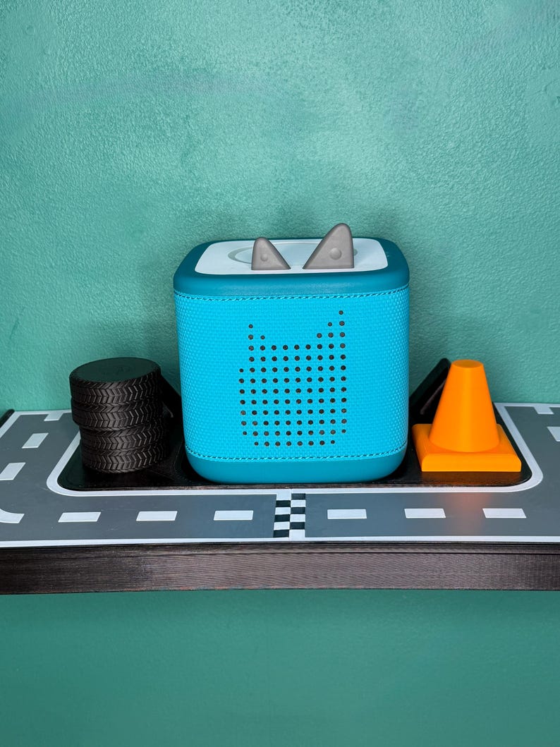 May include: A teal blue audio player with a perforated speaker design and grey accents. The player has a square shape with rounded corners and a white top. A stack of black tyres and an orange traffic cone are also visible.