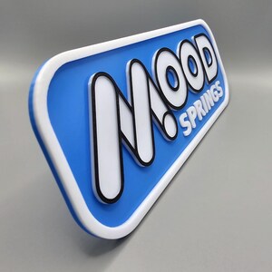 May include: A rectangular sign with rounded corners, featuring the text "MOOD SPRINGS" in white, outlined in black, against a blue background. The sign has a white border and is angled slightly.