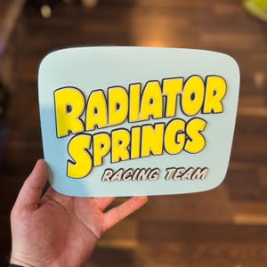 Puede incluir: Un letrero azul claro con las palabras "RADIATOR SPRINGS" en amarillo, delineadas en negro, y "RACING TEAM" en blanco. El letrero tiene esquinas redondeadas y est&aacute; sostenido por una mano.