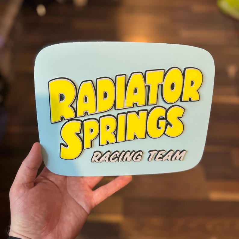 Puede incluir: Un letrero azul claro con las palabras "RADIATOR SPRINGS" en amarillo, delineadas en negro, y "RACING TEAM" en blanco. El letrero tiene esquinas redondeadas y est&aacute; sostenido por una mano.