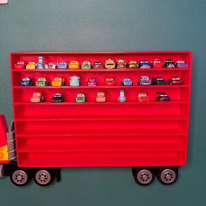 May include: A bright red wall-mounted display case shaped like a semi-truck trailer, featuring multiple horizontal shelves filled with various small, colorful die-cast toy cars with cartoon-style eyes on their windshields, arranged in three rows.