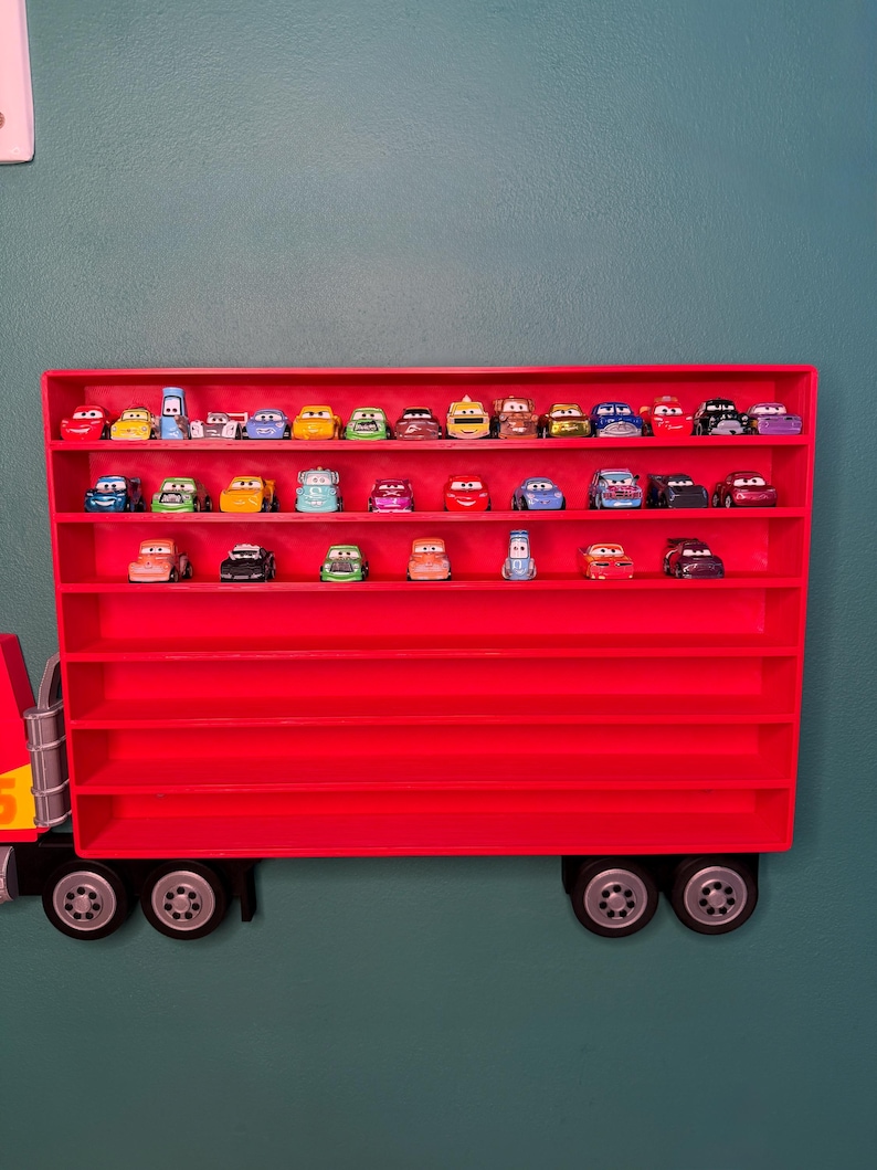 May include: A bright red wall-mounted display case shaped like a semi-truck trailer, featuring multiple horizontal shelves filled with various small, colorful die-cast toy cars with cartoon-style eyes on their windshields, arranged in three rows.