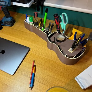 Guitar-shaped desk organiser in use, filled with pens, tools, and supplies. Made from wood-coloured PLA, adding a unique touch to any workspace.