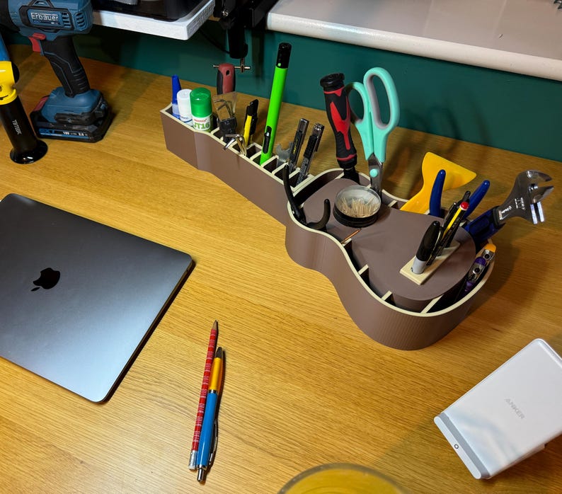 Guitar-shaped desk organiser in use, filled with pens, tools, and supplies. Made from wood-coloured PLA, adding a unique touch to any workspace.