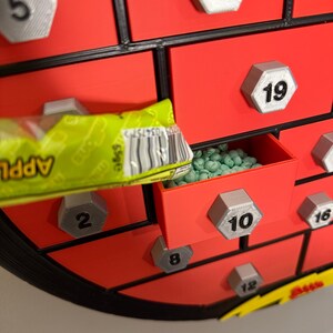 May include: A red advent calendar with multiple small drawers, each labeled with a number. One drawer is open, revealing green candies. A green and yellow candy wrapper is partially visible, with the word "APPLE" printed on it.