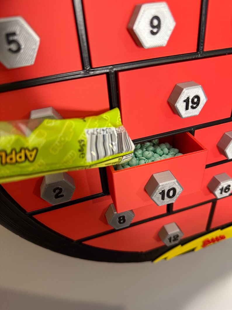 May include: A red advent calendar with multiple small drawers, each labeled with a number. One drawer is open, revealing green candies. A green and yellow candy wrapper is partially visible, with the word "APPLE" printed on it.