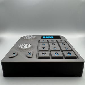 May include: A gray rectangular device with a black base, featuring a keypad with blue illuminated numbers and the text "HARV". The device has speaker and button details. The overall design is modern and functional.
