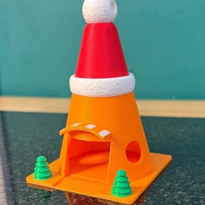 Può includere: Una struttura a forma di cono stradale arancione sormontata da un cappello da Babbo Natale rosso e bianco. La base presenta due piccoli pini verdi ai lati di un ingresso con una piccola tettoia e un'apertura laterale circolare.