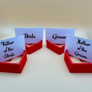 Set of 3D-printed red heart-shaped place card holders showcasing “Bride,” “Groom,” “Mother of the Groom,” and “Father of the Bride” cards. Perfect for wedding table décor.  Durable and stylish, they enhance romantic and elegant table settings.