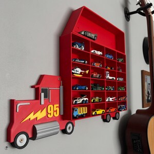 May include: Red toy truck with a number 95 on the side, attached to a red wall shelf with multiple slots for toy cars. The shelf is shaped like a barn with a red roof.