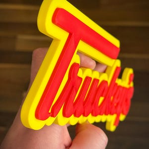 K&ouml;nnte beinhalten: Ein gelbes und rotes Schild mit dem Wort "Truckers" in einer stilisierten Schriftart. Das Schild wird in einer Hand gehalten und hat ein dreidimensionales Aussehen. Der Hintergrund ist eine verschwommene Holzoberfl&auml;che.