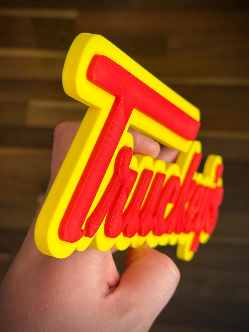 K&ouml;nnte beinhalten: Ein gelbes und rotes Schild mit dem Wort "Truckers" in einer stilisierten Schriftart. Das Schild wird in einer Hand gehalten und hat ein dreidimensionales Aussehen. Der Hintergrund ist eine verschwommene Holzoberfl&auml;che.