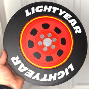 May include: A black and red decorative wheel with the word "LIGHTYEAR" in white lettering. The wheel has a red center with silver accents and a yellow stripe. The design resembles a car tire.