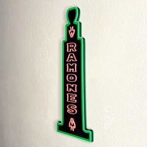 Puede incluir: Un letrero negro y verde con la palabra "RAMONES" en letras rosas. El letrero tiene una forma &uacute;nica con un contorno verde y est&aacute; montado en una pared de color claro. El letrero tiene una llama y un s&iacute;mbolo en la parte superior e inferior.