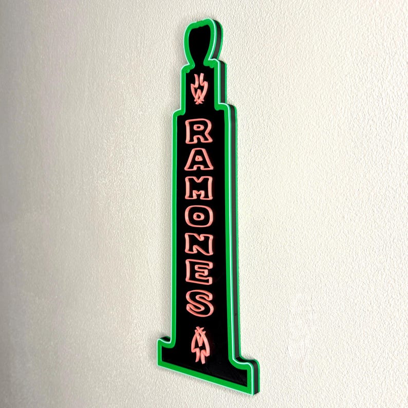 Puede incluir: Un letrero negro y verde con la palabra "RAMONES" en letras rosas. El letrero tiene una forma &uacute;nica con un contorno verde y est&aacute; montado en una pared de color claro. El letrero tiene una llama y un s&iacute;mbolo en la parte superior e inferior.