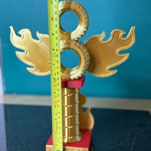 May include: A gold-coloured trophy with a flame design and two circular elements. The trophy is approximately 28 cm tall, with a red base and the word "NER" visible. The design includes wing-like shapes and a cylindrical column.