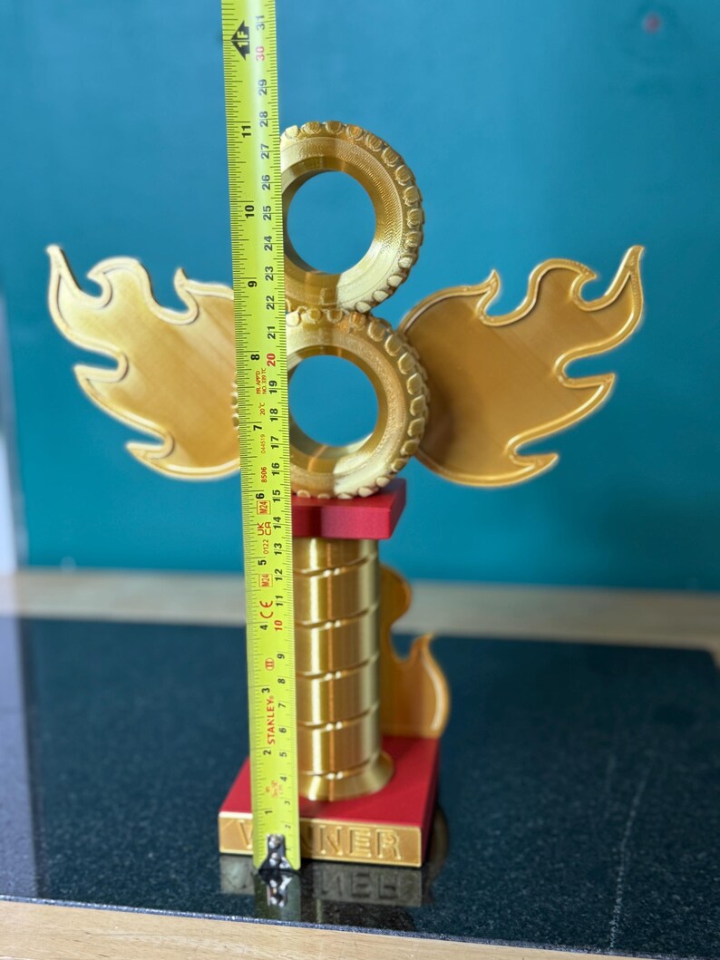 May include: A gold-coloured trophy with a flame design and two circular elements. The trophy is approximately 28 cm tall, with a red base and the word "NER" visible. The design includes wing-like shapes and a cylindrical column.