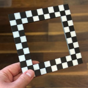 May include: A square picture frame with a black and white checkered pattern. The frame is made of a dark material and features alternating black and white squares. The frame is held by a hand.