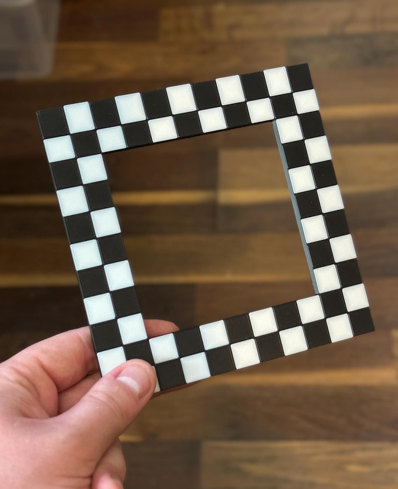 May include: A square picture frame with a black and white checkered pattern. The frame is made of a dark material and features alternating black and white squares. The frame is held by a hand.