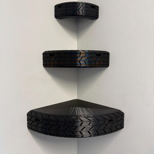 May include: Three black corner shelves with a tire tread design are mounted on a white wall. The shelves are triangular and semi-circular, with the largest at the bottom. The shelves are empty.