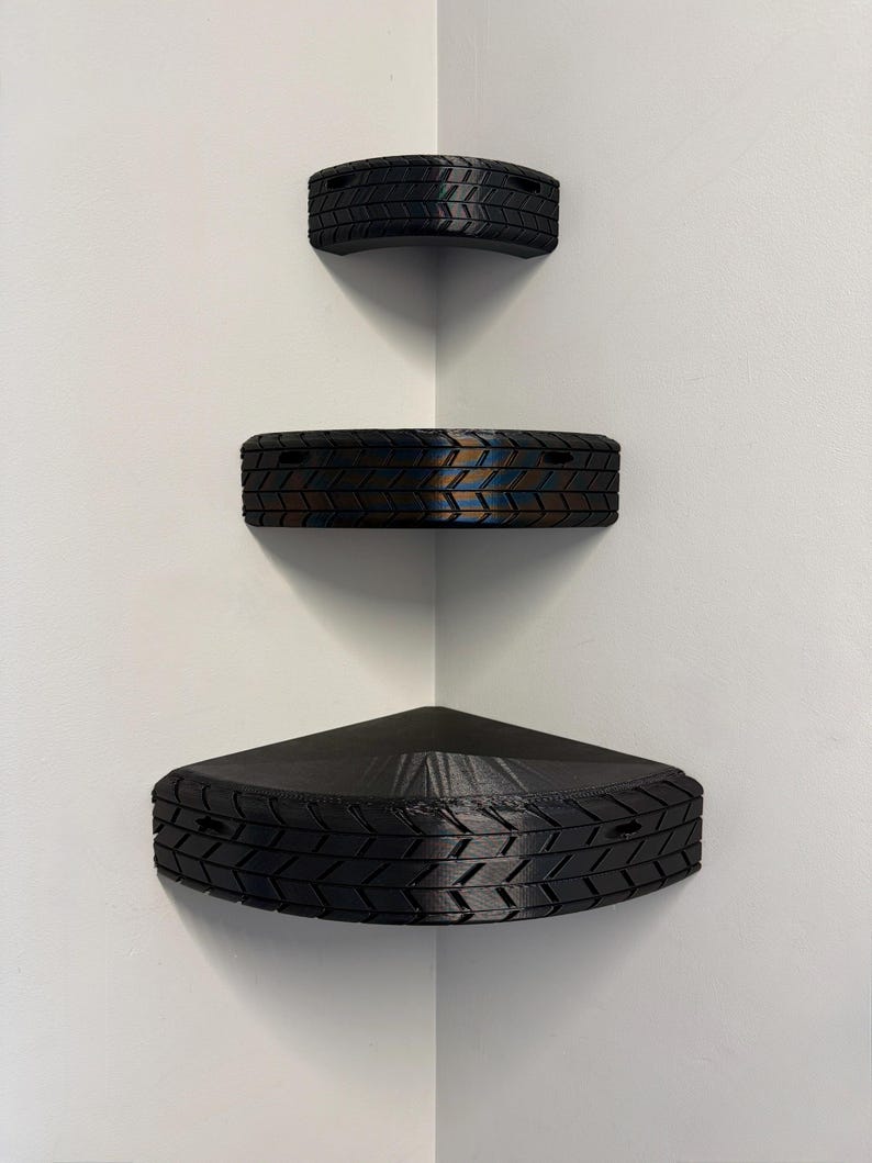 May include: Three black corner shelves with a tire tread design are mounted on a white wall. The shelves are triangular and semi-circular, with the largest at the bottom. The shelves are empty.