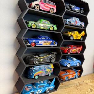 May include: A black honeycomb-shaped wall display shelf featuring multiple hexagonal compartments. Each individual slot contains a miniature toy car, including various colorful character vehicles from the Cars movie franchise, arranged in a vertical grid.
