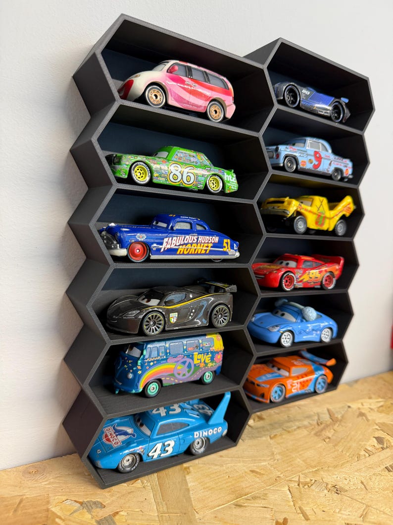 May include: A black honeycomb-shaped wall display shelf featuring multiple hexagonal compartments. Each individual slot contains a miniature toy car, including various colorful character vehicles from the Cars movie franchise, arranged in a vertical grid.