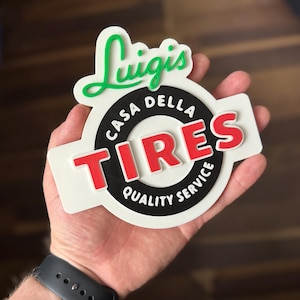 May include: A white, black, and red sign with the text "Luigi's CASA DELLA TIRES QUALITY SERVICE". The sign has a green, cursive "Luigi's" at the top. The word "TIRES" is in red, and the rest of the text is in white.