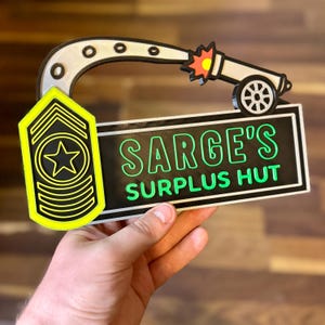 May include: A novelty sign featuring a cartoon cannon firing a projectile. The sign includes the text "SARGE'S SURPLUS HUT" in green lettering on a black background. A yellow and black military rank insignia is also present.
