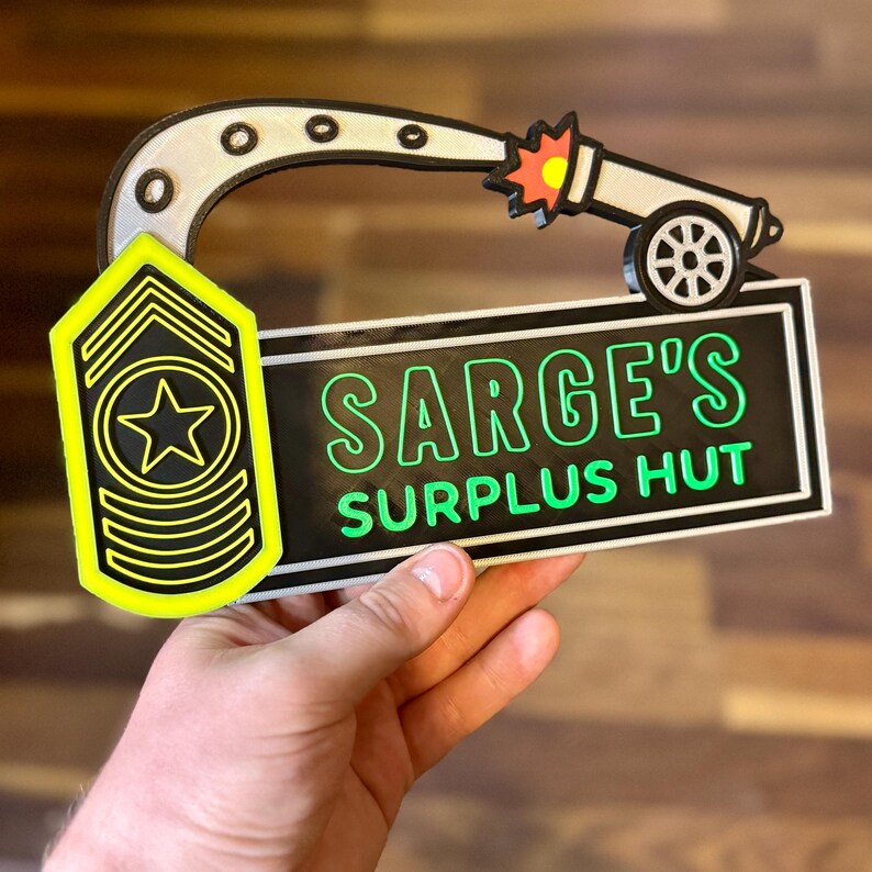 May include: A novelty sign featuring a cartoon cannon firing a projectile. The sign includes the text "SARGE'S SURPLUS HUT" in green lettering on a black background. A yellow and black military rank insignia is also present.