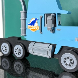 May include: A light blue toy truck with the Dinoco logo on the side. The truck has a black chassis, gray wheels, and a silver exhaust pipe. The Dinoco logo features a blue dinosaur silhouette.