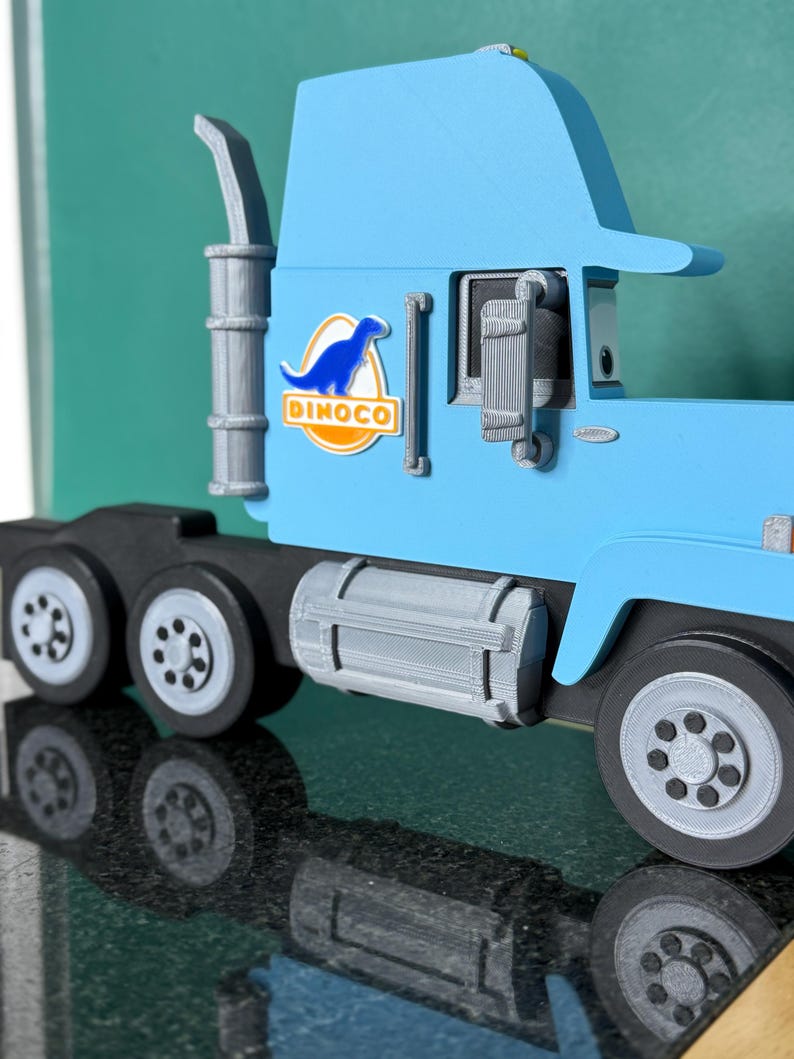 May include: A light blue toy truck with the Dinoco logo on the side. The truck has a black chassis, gray wheels, and a silver exhaust pipe. The Dinoco logo features a blue dinosaur silhouette.