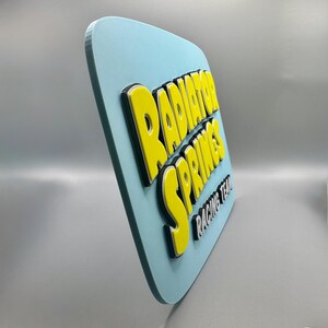 Puede incluir: Un letrero azul claro con esquinas redondeadas presenta las palabras "RADIATOR SPRINGS" en amarillo, delineadas en negro, y "RACING TEAM" en blanco. El letrero tiene un efecto 3D.