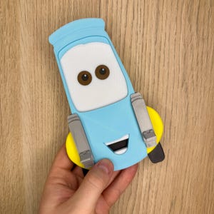 May include: A light blue, white, and brown cartoon car character with a smiling face. The character has brown eyes, a white windshield, and gray side panels. The car has a yellow bumper and a black tire.