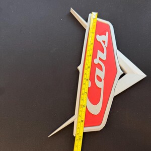 May include: A red and white Cars logo with a silver outline, measured with a yellow tape measure. The logo is approximately 11 inches in length.