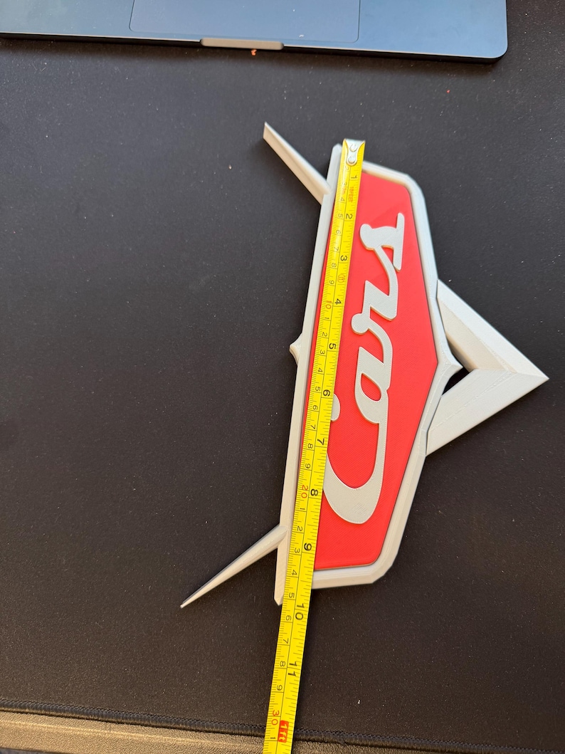 May include: A red and white Cars logo with a silver outline, measured with a yellow tape measure. The logo is approximately 11 inches in length.